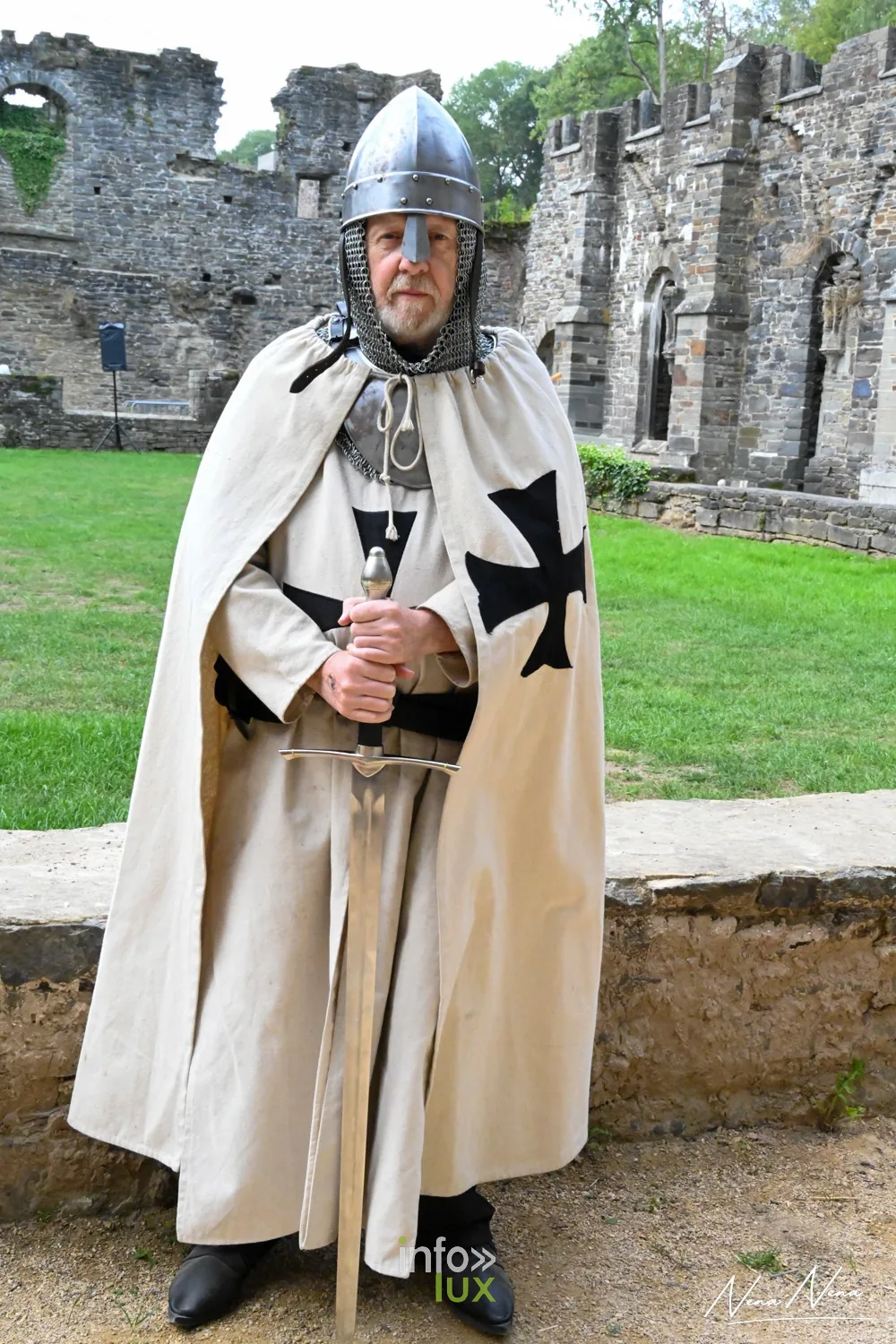 Un chevalier en cape blanche à croix noire tient son épée devant lui, dans les ruines