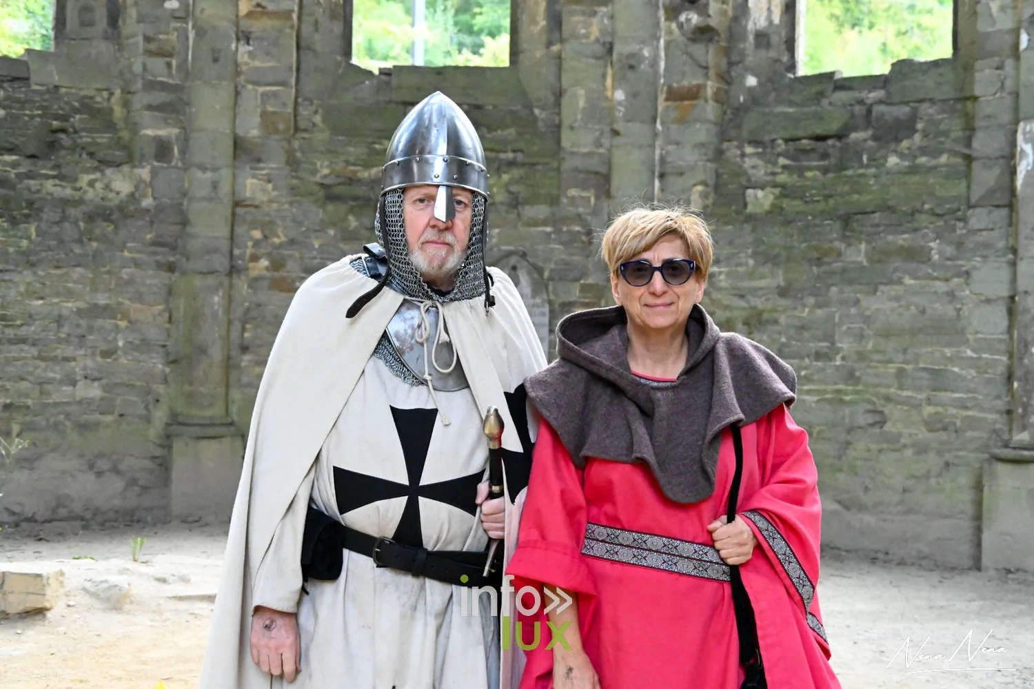Un chevalier en cape blanche pose avec une visiteuse en robe rouge devant les ruines