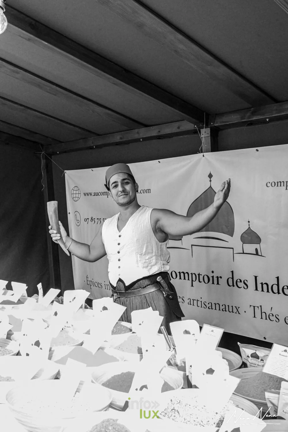 Un marchand d’épices en gilet blanc, bras ouverts devant sa bannière « Au Comptoir des Indes », en noir et blanc