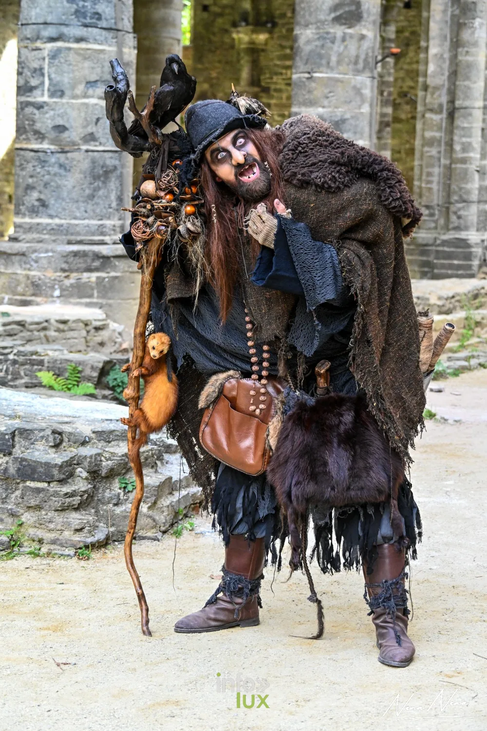 Un sorcier en haillons et fourrures, corbeau sur l’épaule et bâton en main, dans les ruines