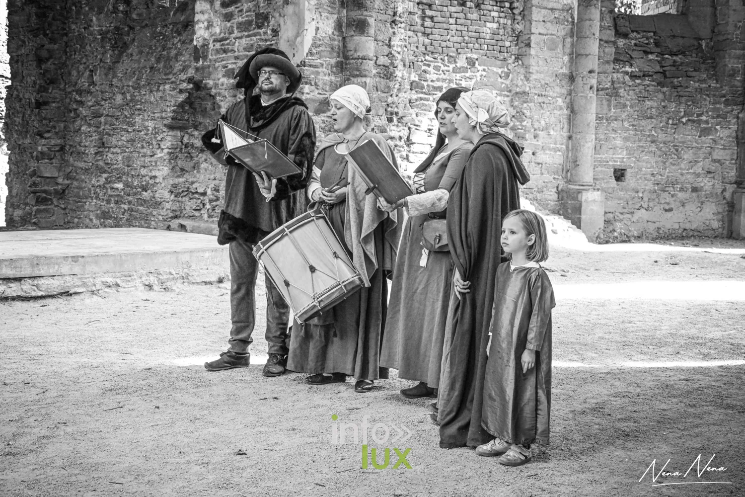 Un groupe de musiciens en costume, tambour et livrets de chant, sous les arches de l’abbatiale, en noir et blanc
