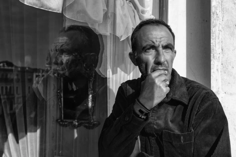 Portrait en noir et blanc de Nikos Aliagas devant une fenetre, son reflet dans la vitre