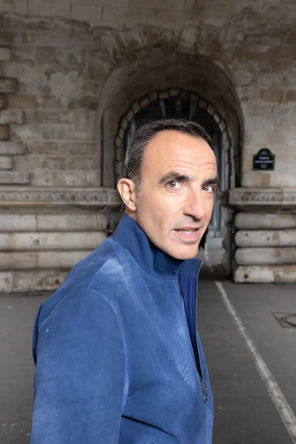 Nikos Aliagas de trois quarts sous une arche de pierre a Paris