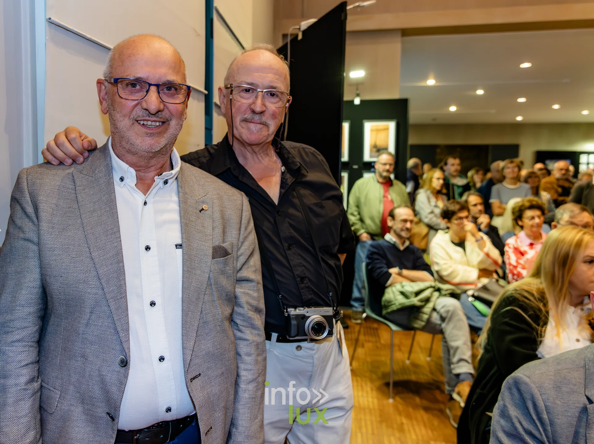 Deux invités posant devant la salle lors du vernissage du Royal Photo Club Entre Nous, l’un avec un appareil photo