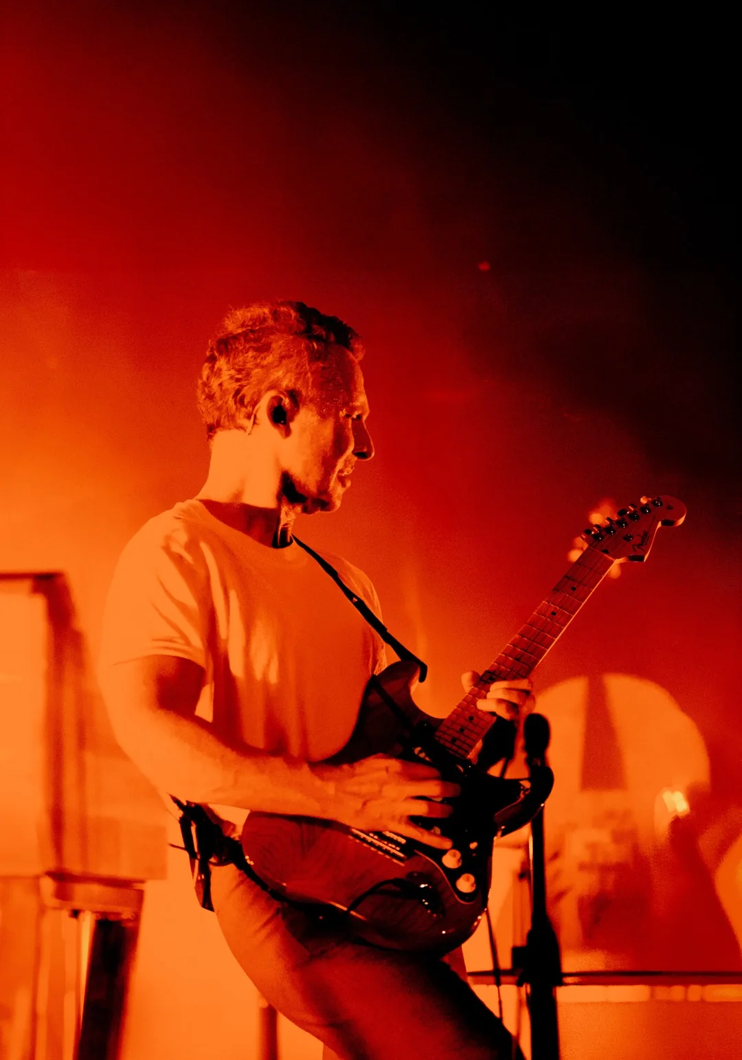 Sinclair de profil, guitare en bandoulière, dans une lumière orange et rouge