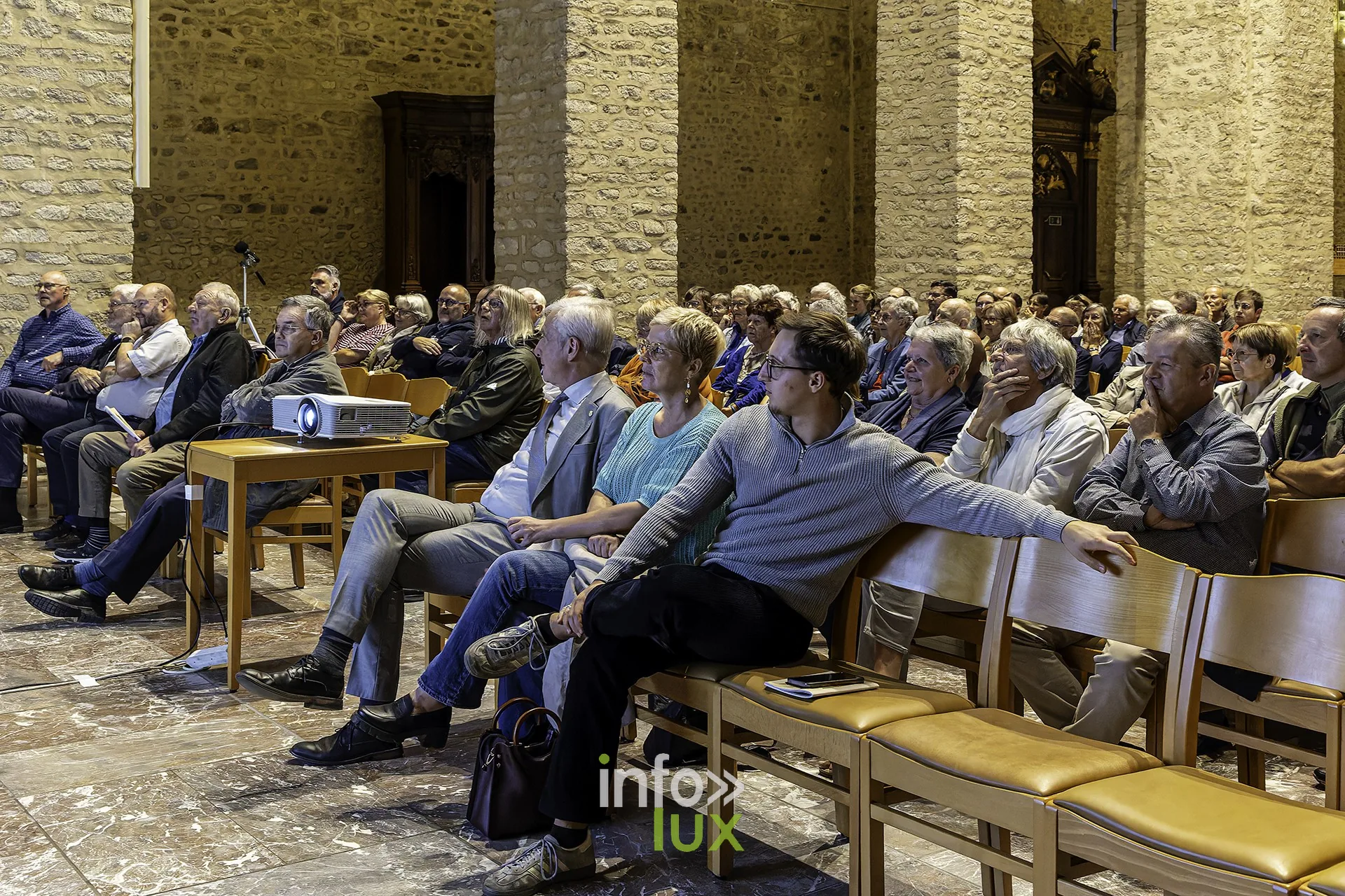 Rangées de spectateurs assis, de profil, dans la lumière des vitraux