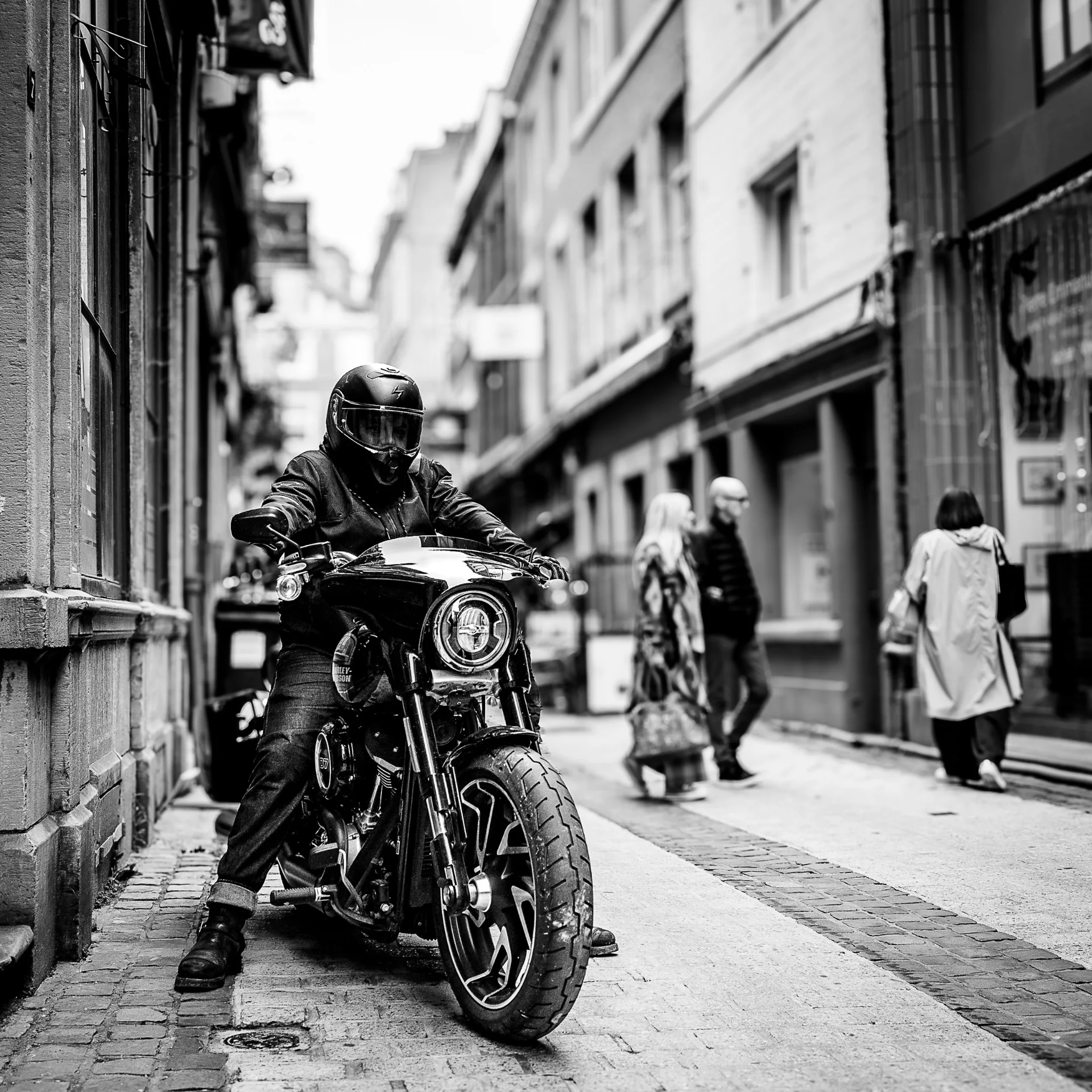 Un motard casqué avance dans une ruelle pavée de Liège au milieu des passants, photo de rue en noir et blanc