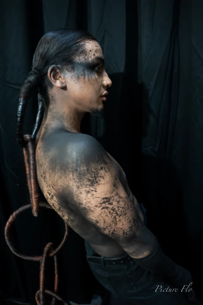 Youness de profil en coulisses, natte et anneaux en cheveux, épaule maquillée de noir, Lux Fashion Week 2026, Arlon
