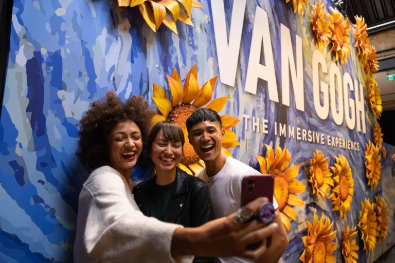 Trois visiteurs font un selfie devant le mur aux tournesols de Van Gogh: The Immersive Experience