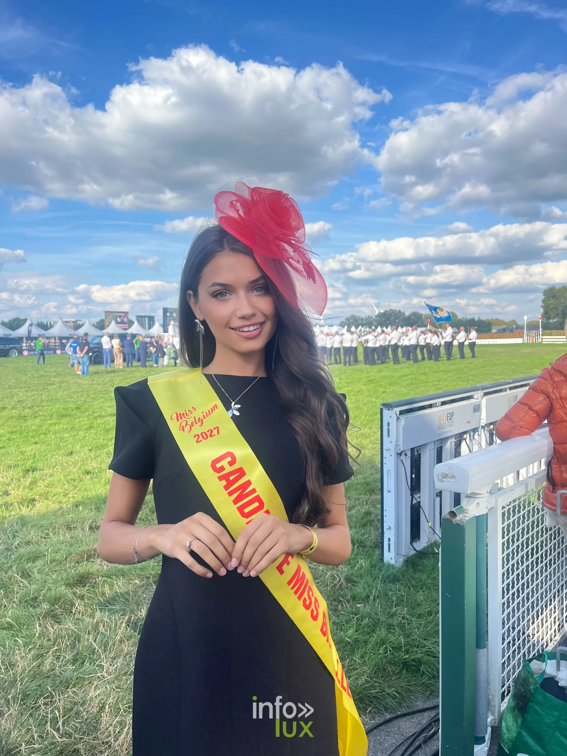 Elisa en écharpe de candidate Miss Belgique 2027 et bibi rouge, lors d’un événement en plein air