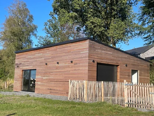 Un bâtiment au bardage de bois et toit plat, entouré de pelouse et d’une clôture en ganivelle