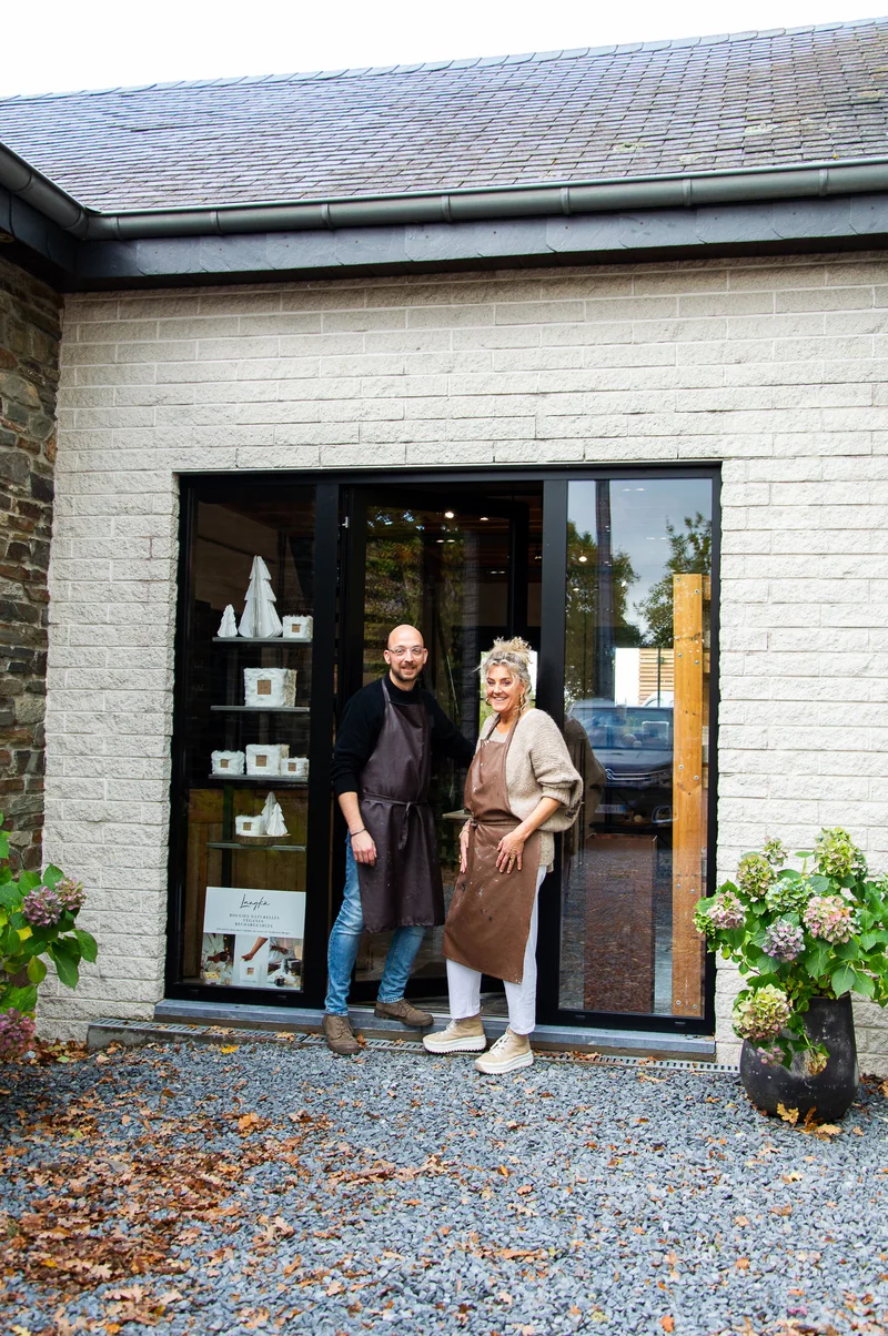 Une femme et un homme en tablier brun posent sur le seuil d’une boutique Langkâ aux vitrines garnies de bougies