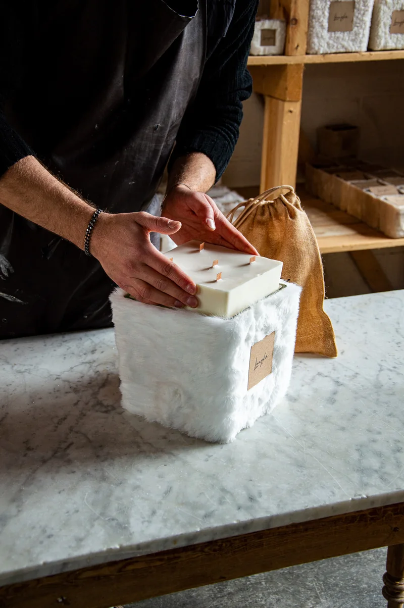 Des mains posent une bougie blanche à plusieurs mèches dans un contenant habillé de fausse fourrure blanche