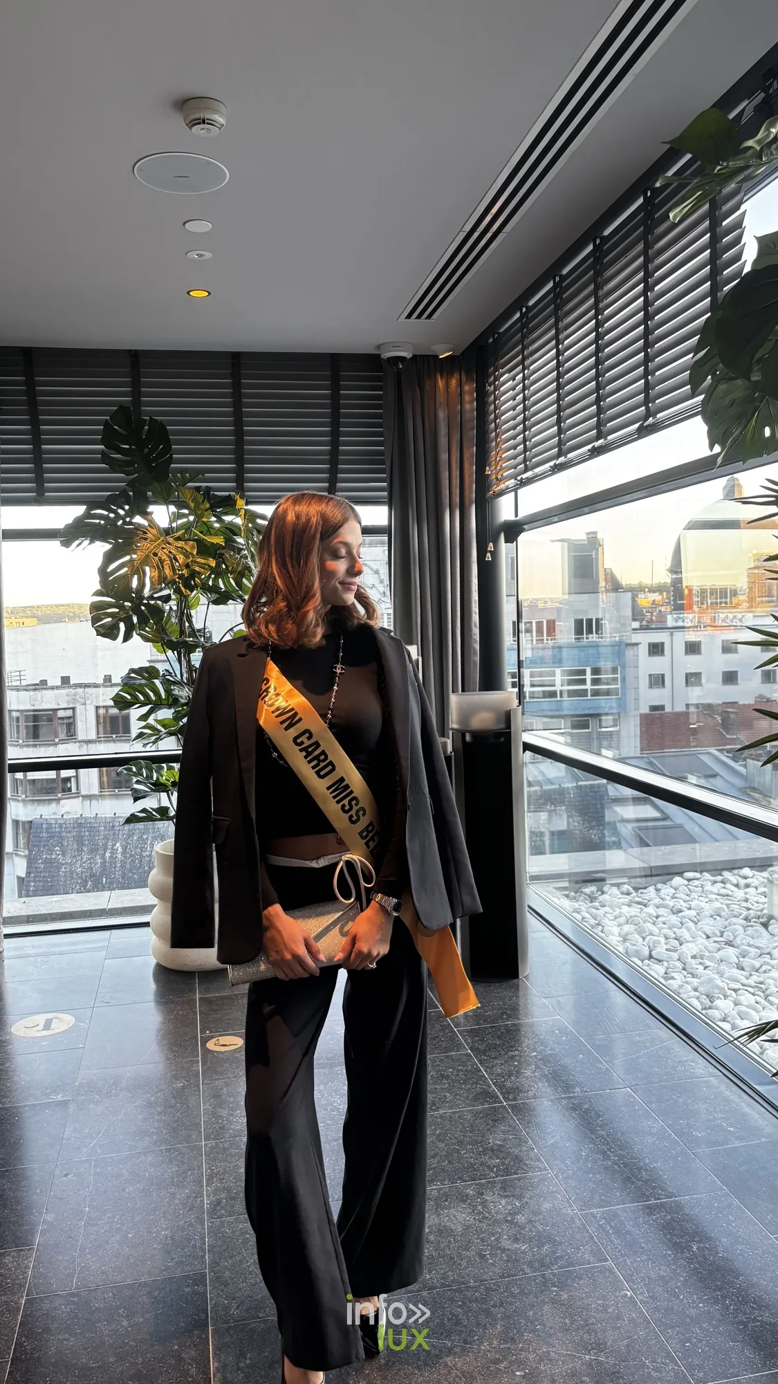 Lauren en tailleur noir avec son écharpe Crown Card Miss Belgium, dans un espace vitré avec vue sur la ville