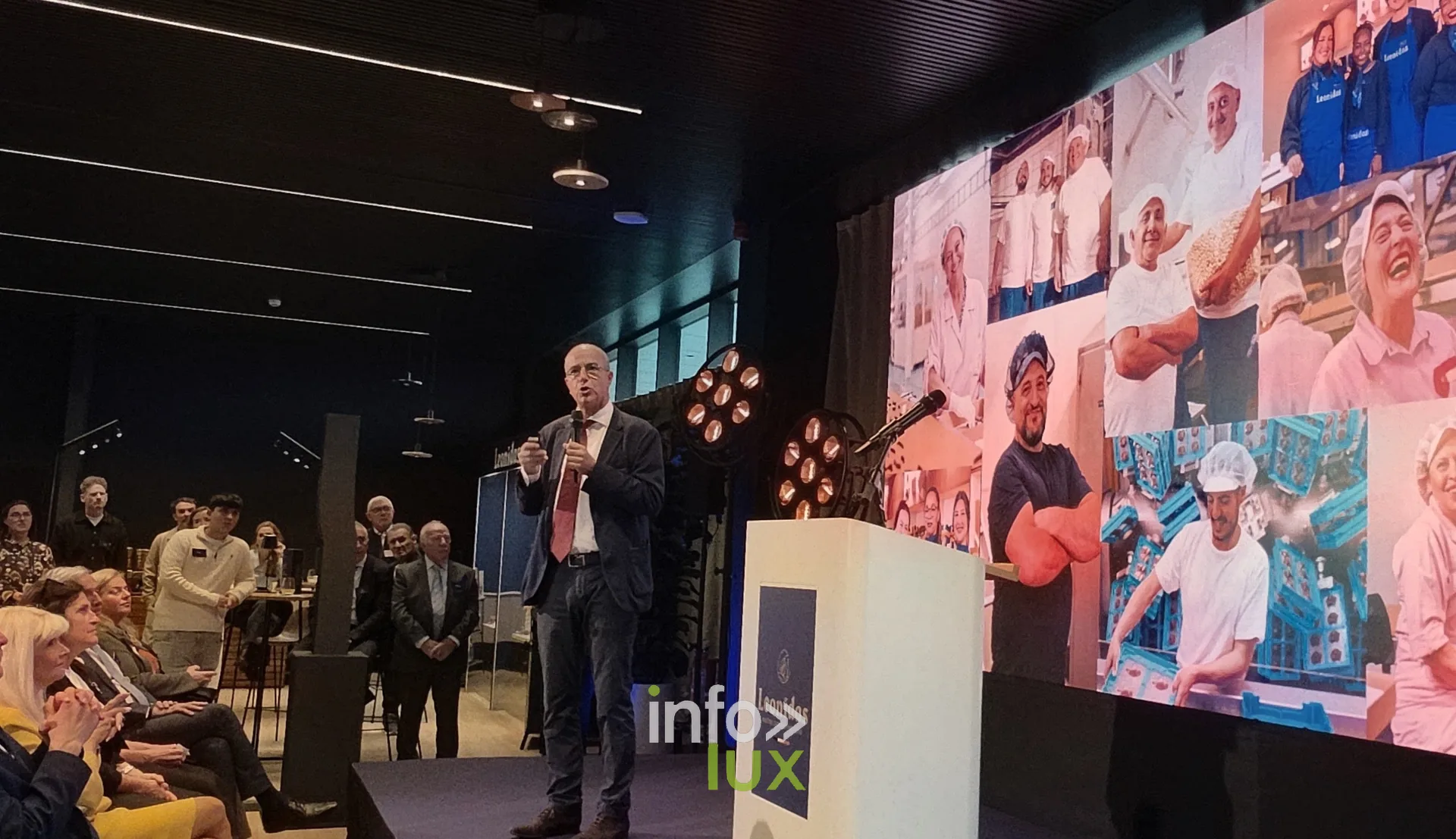 Un discours devant un écran montrant des photos d’équipes au travail, inauguration du nouveau site Leonidas à Nivelles, 15 octobre 2026 (photo Karolyn Delisé-Hanse) — photo modifiée par ia