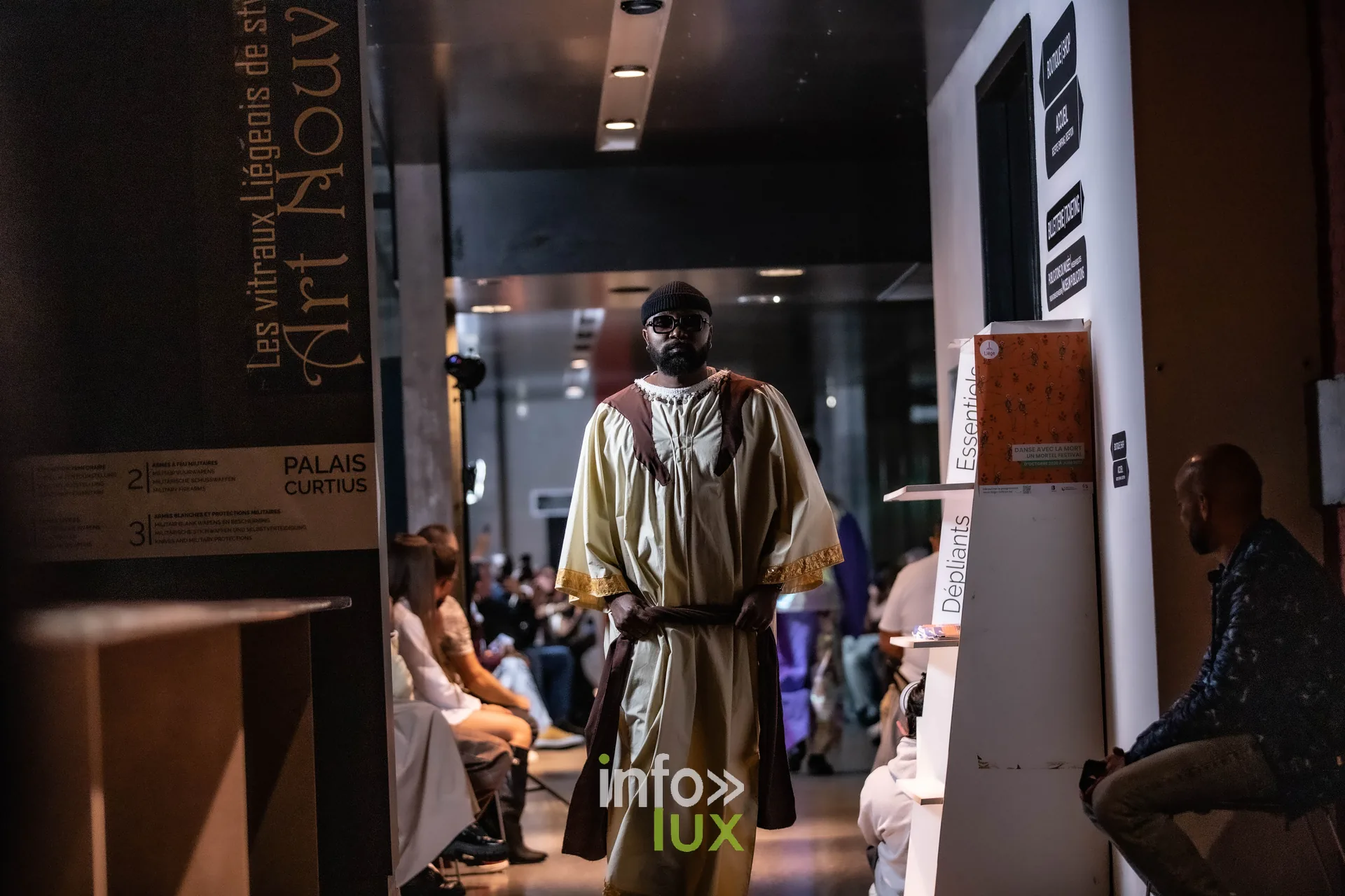 Liège Fashion Week 2026 au Musée Grand Curtius à Liège : mannequin masculin en tunique beige et ceinture marron, panneau Palais Curtius