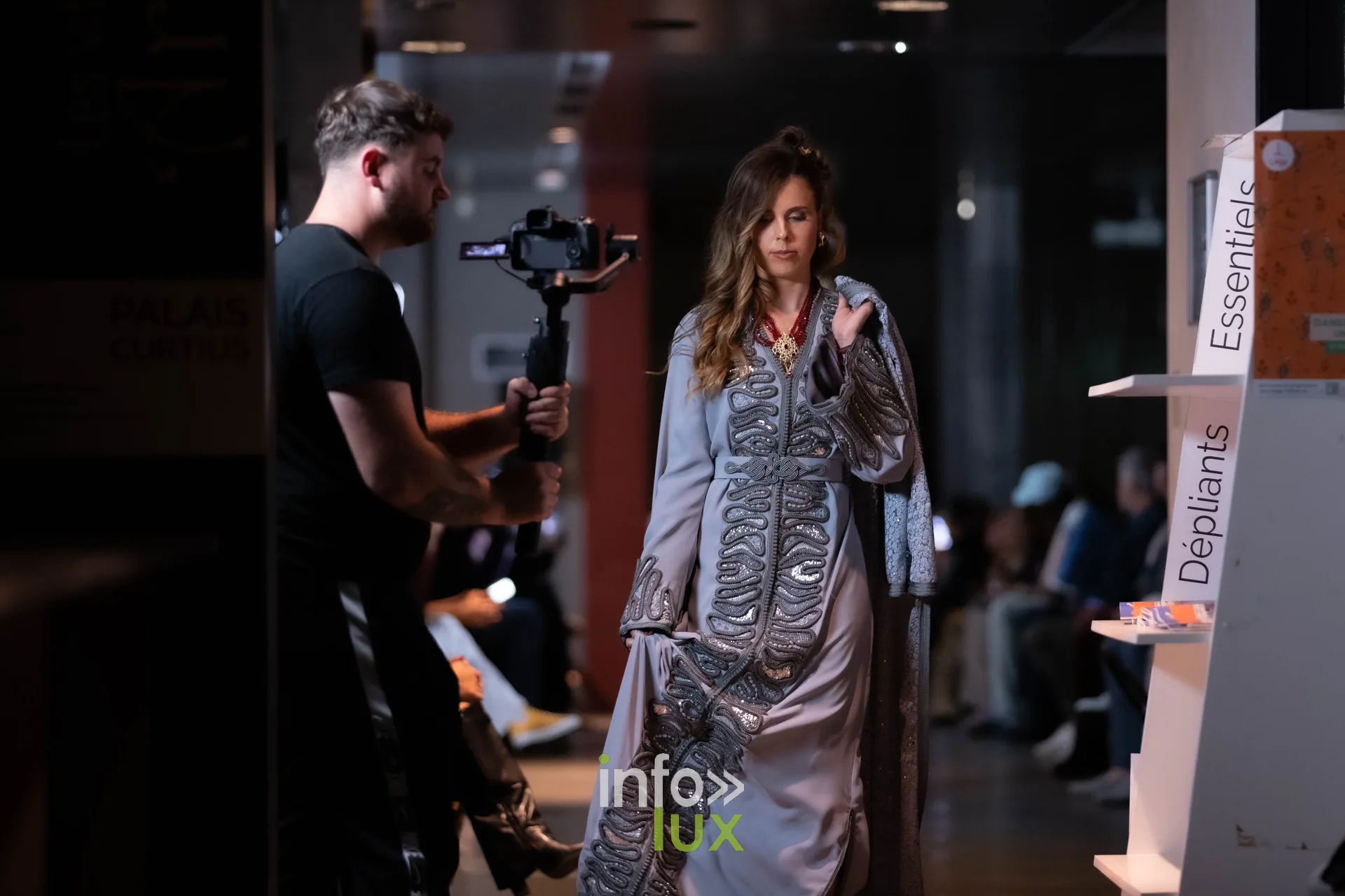 Liège Fashion Week 2026 au Musée Grand Curtius à Liège : mannequin en caftan gris argent brodé, suivie par un caméraman