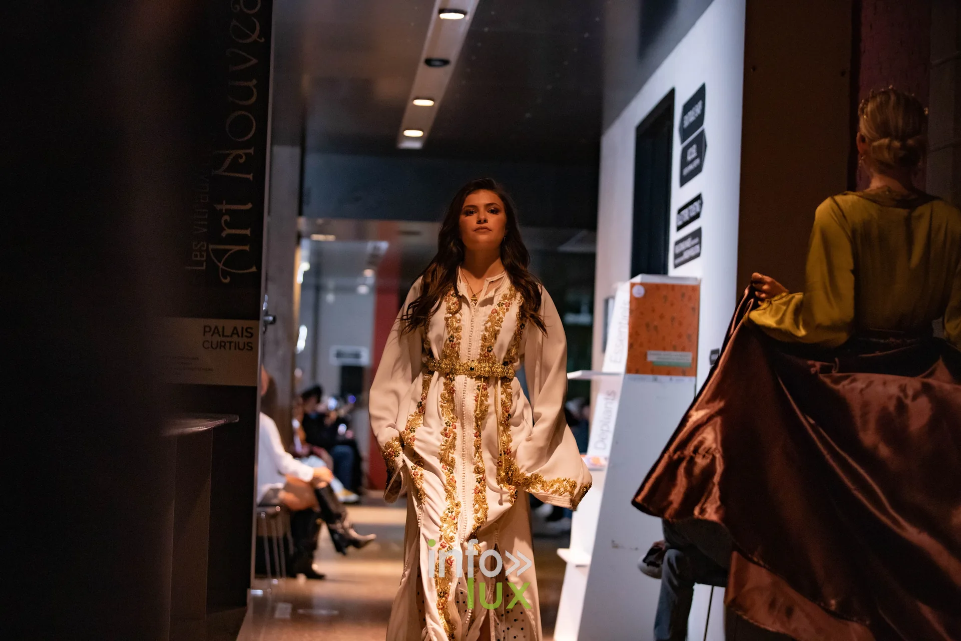 Liège Fashion Week 2026 au Musée Grand Curtius à Liège : mannequin en caftan blanc et or, robe de satin brun au premier plan
