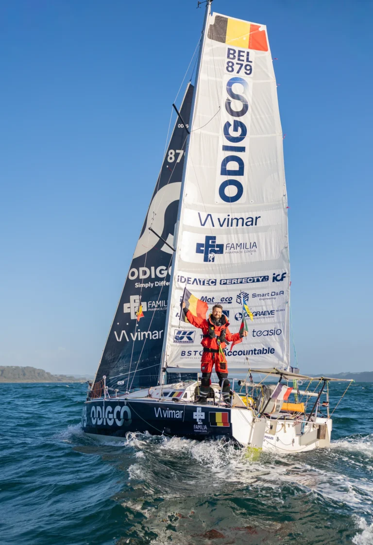 Quentin Debois debout sur son Mini 6.50 ODIGO, drapeau belge en main, à l’arrivée au Cap Lizard