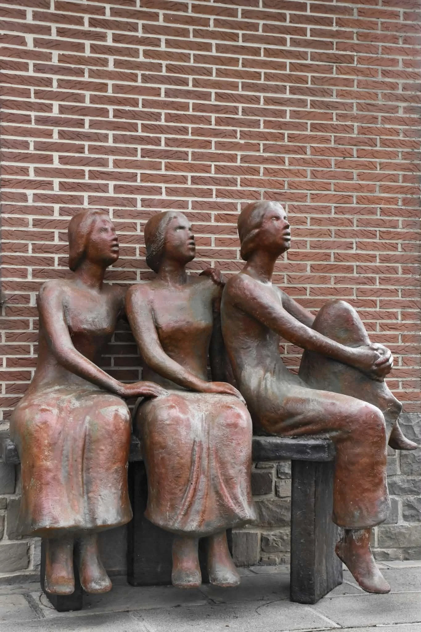 Sculpture de trois femmes assises côte à côte sur un banc, devant un mur de briques