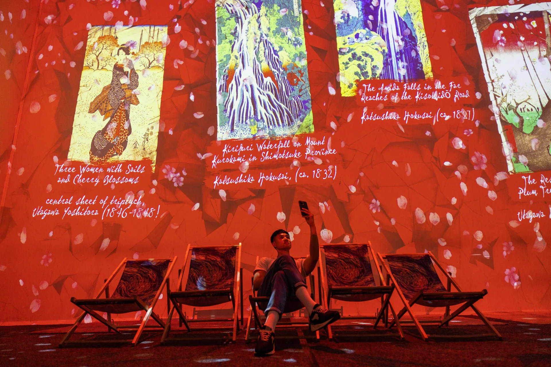 Un visiteur assis dans un transat devant des estampes japonaises de Hokusai et Utagawa Yoshitora projetées sur fond rouge
