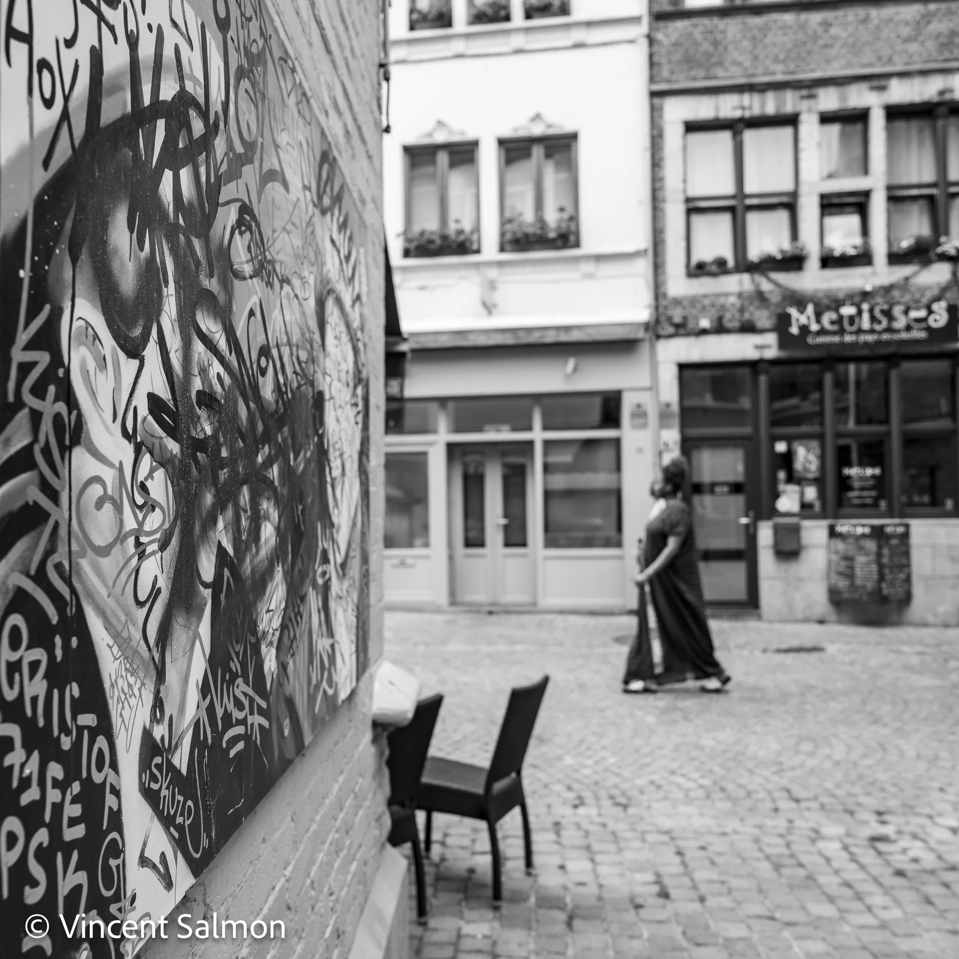 Photo de Vincent Salmon : un mur couvert de graffitis et une passante dans une rue pavée