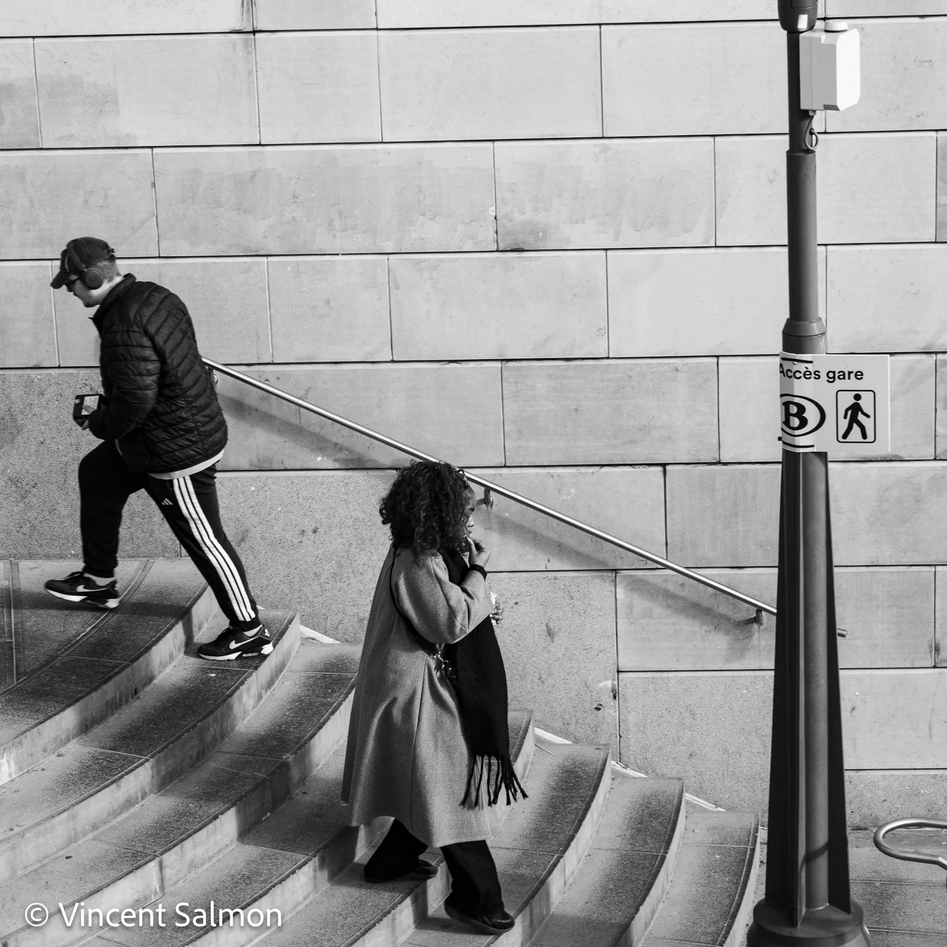 Photo de Vincent Salmon : deux passants dans un escalier près d’un panneau vers la gare