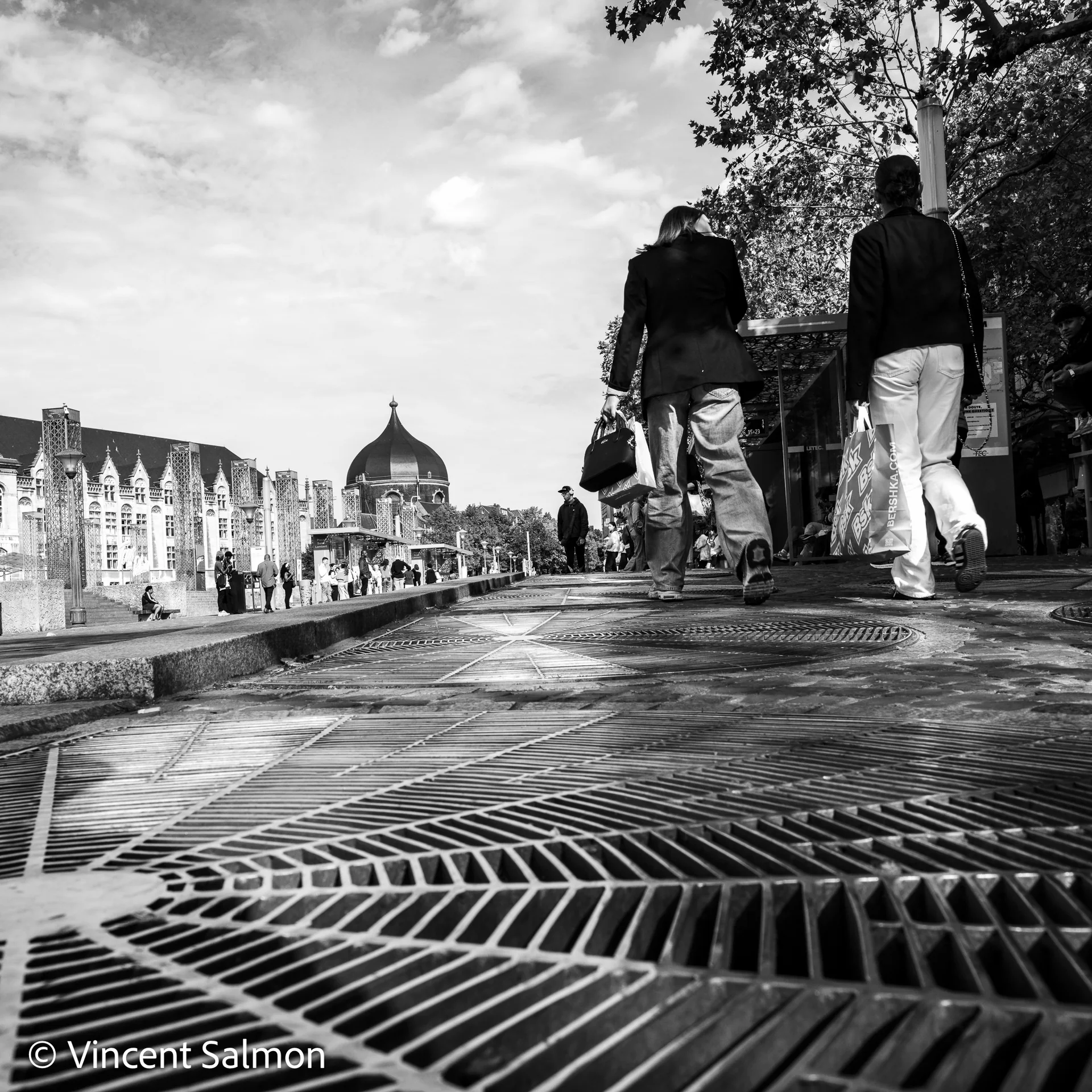 Photo de Vincent Salmon : deux passants au bord d’une place aux grilles mouillées