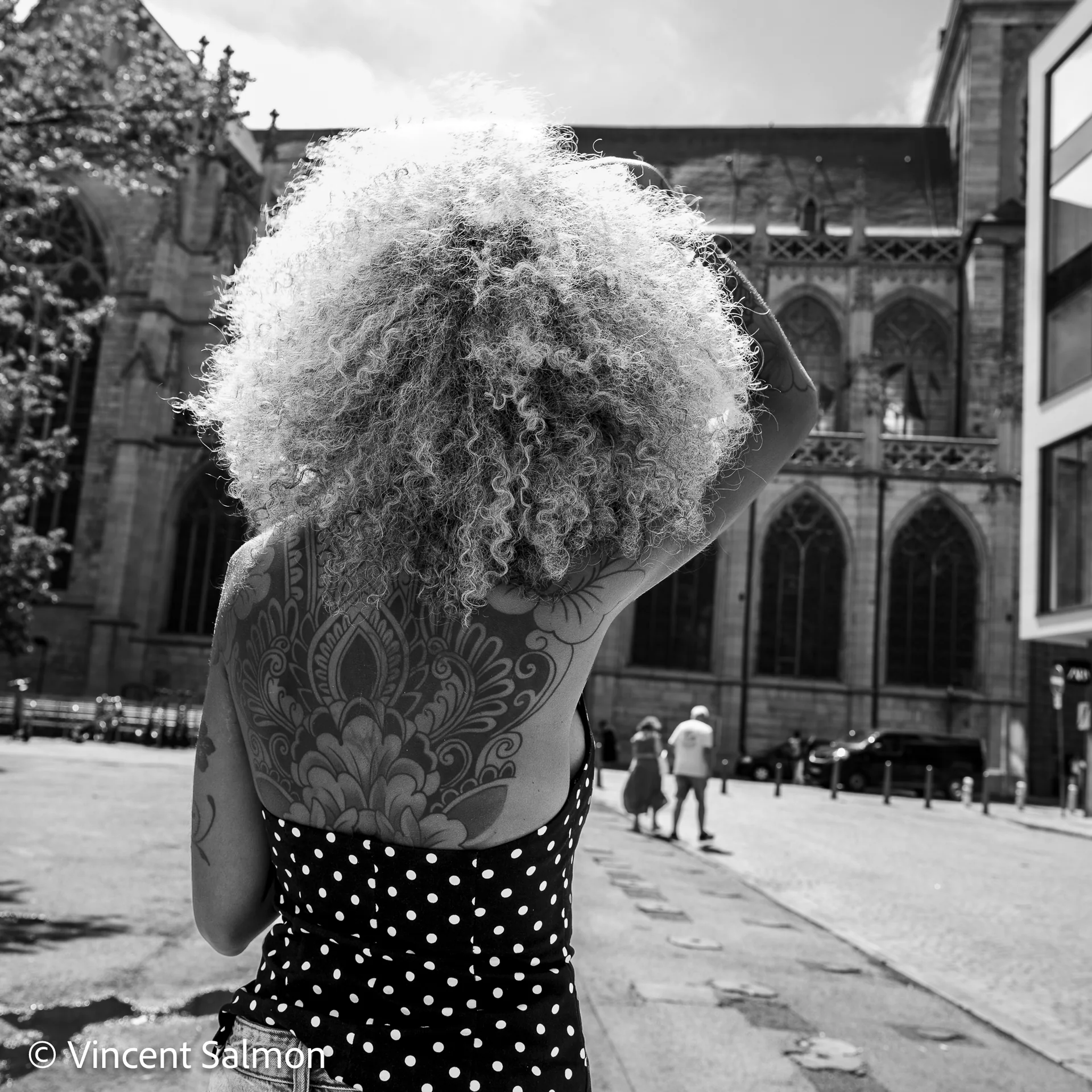 Photo de Vincent Salmon : une femme aux cheveux bouclés et au dos tatoué devant une église